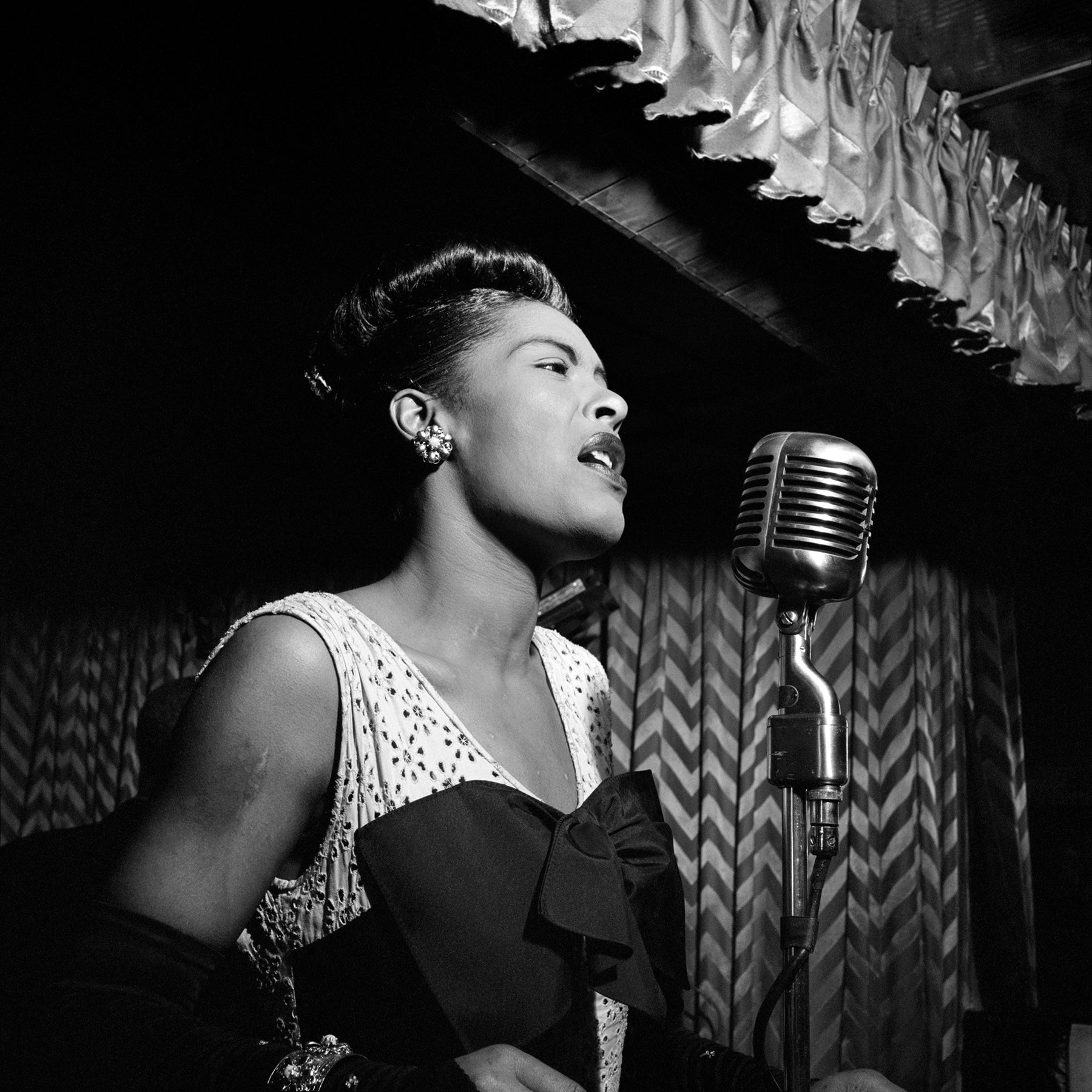 A vintage photograph of Billie Holiday singing into a microphone on stage