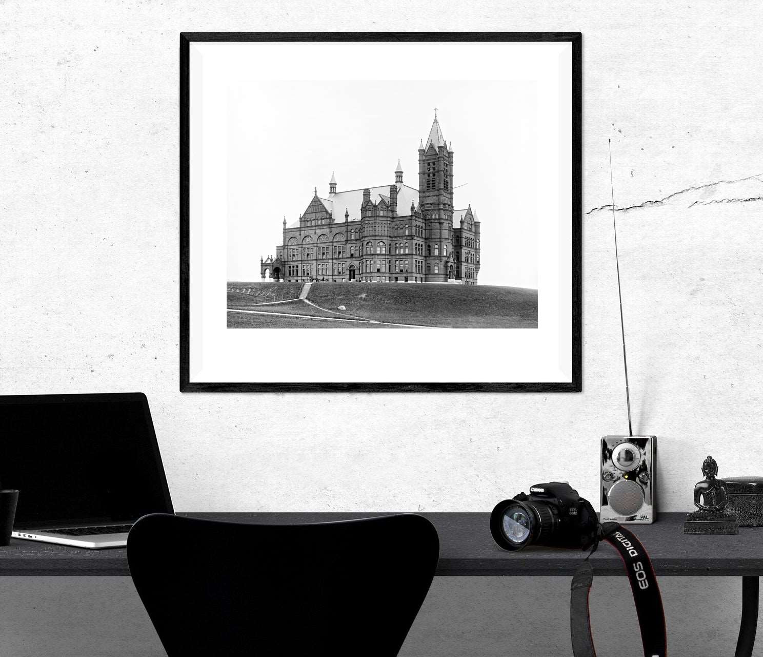 A photograph of a desk with a framed paper print hanging above, featuring a photograph of Syracuse University