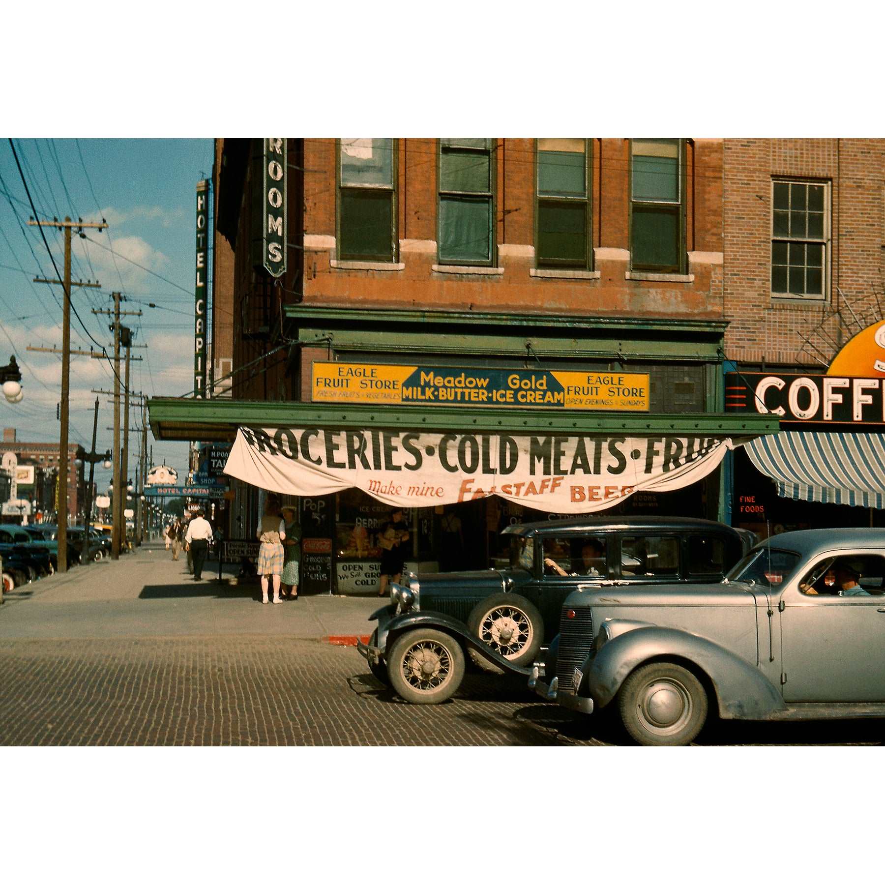 A full color vintage photograph of a fruit store and hotel with cars parked outside