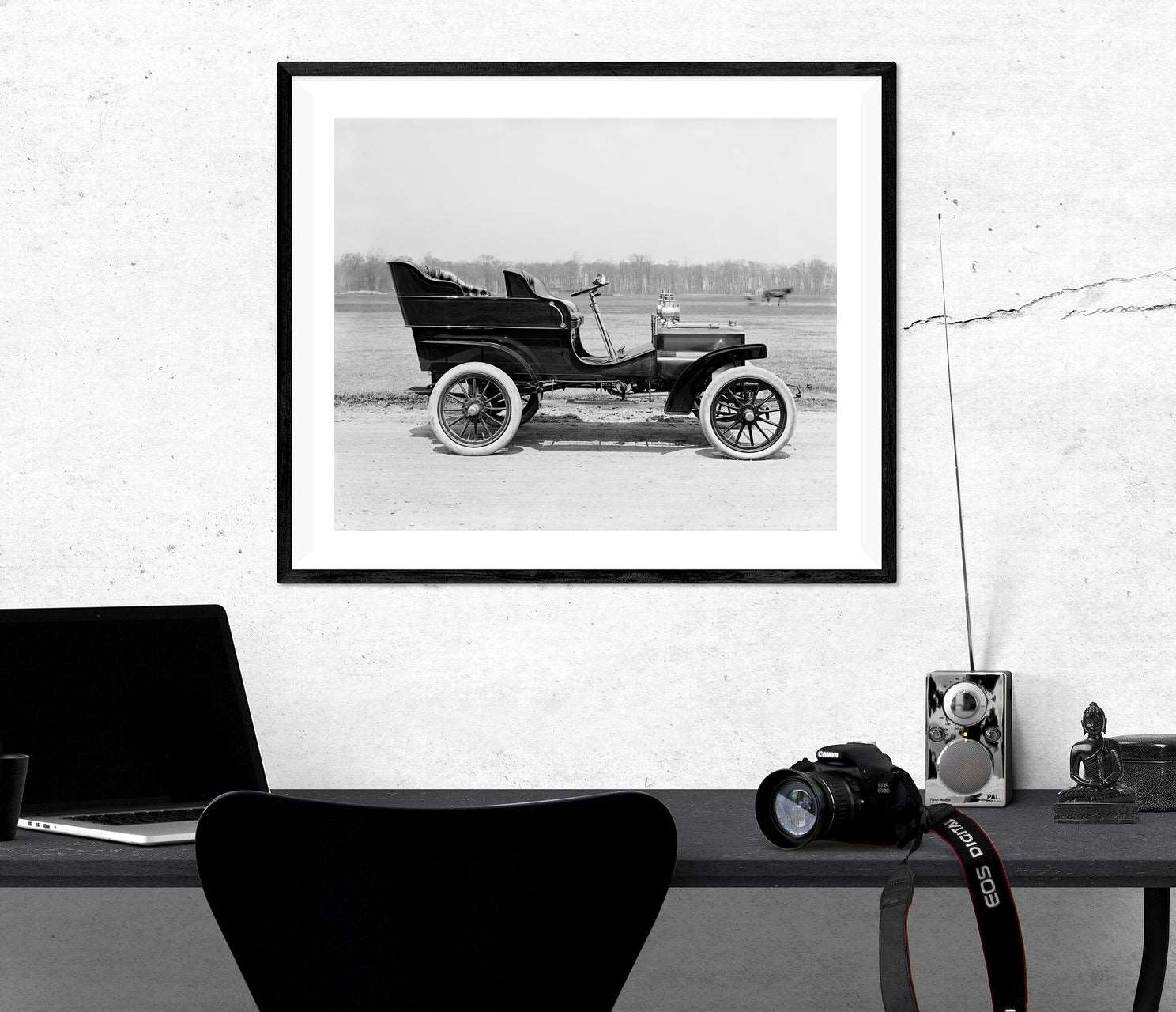 A framed paper print hanging on a white wall, featuring a black and white photo of a vintage vehicle