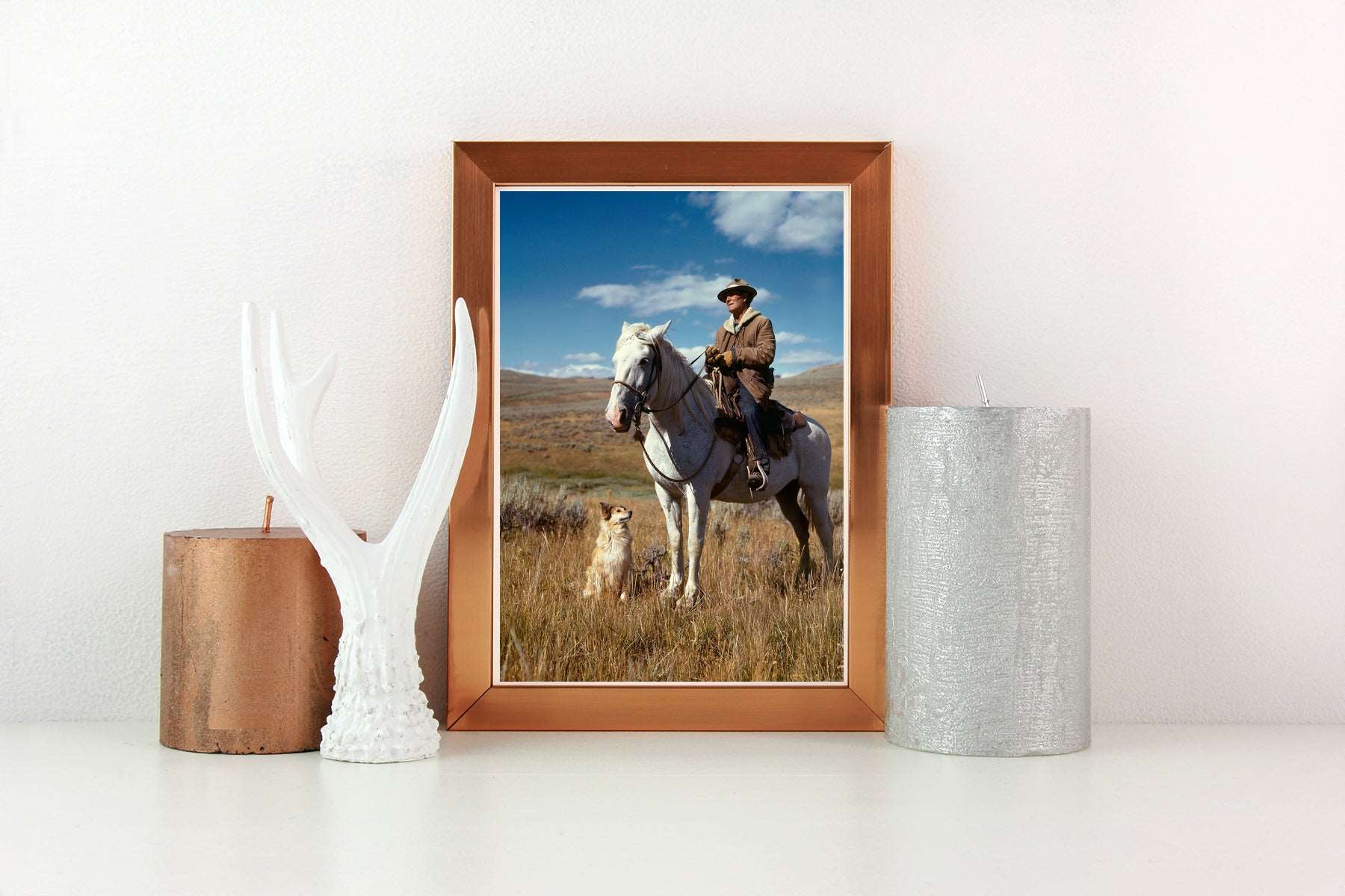 A framed paper print of a color photograph of a shepherd on a horse, displayed on a table with candles