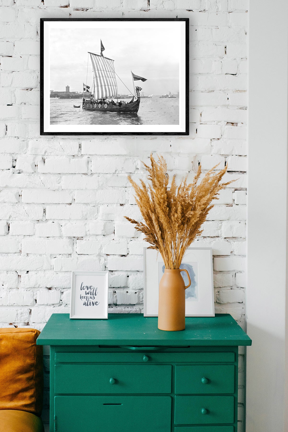 A framed paper print of a vintage photograph hanging on a brick wall above a green set of drawers