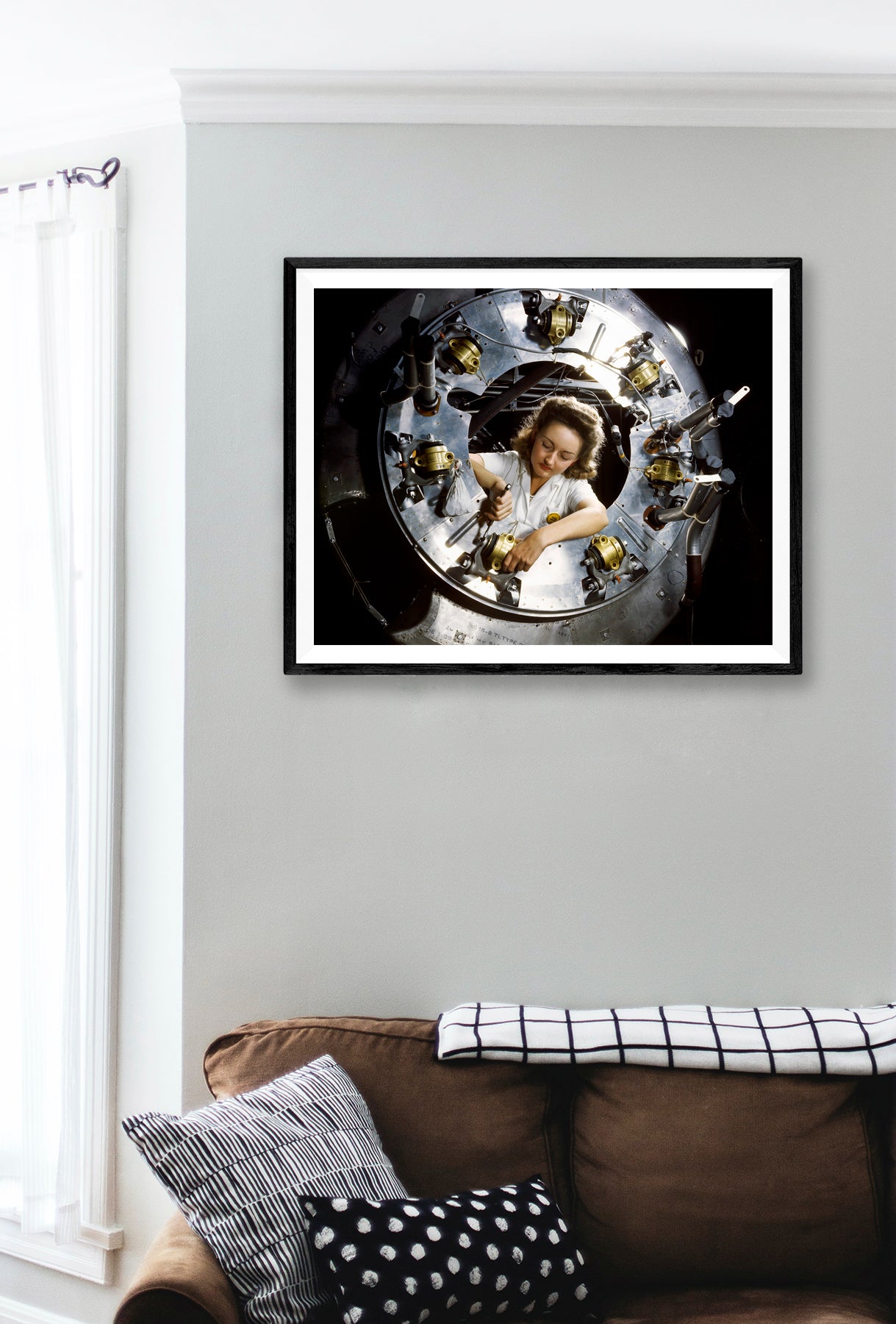 A digital rendering showing a framed paper print of a photograph of a woman working on a motor part of a plane, hanging on a wall above a couch