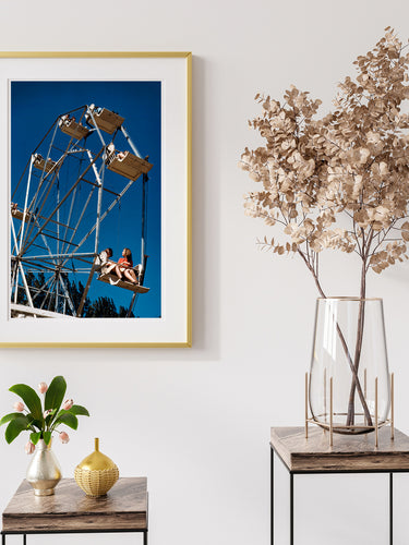 A framed print of a vintage photograph of a ferris wheel hanging on a wall above two small display tables