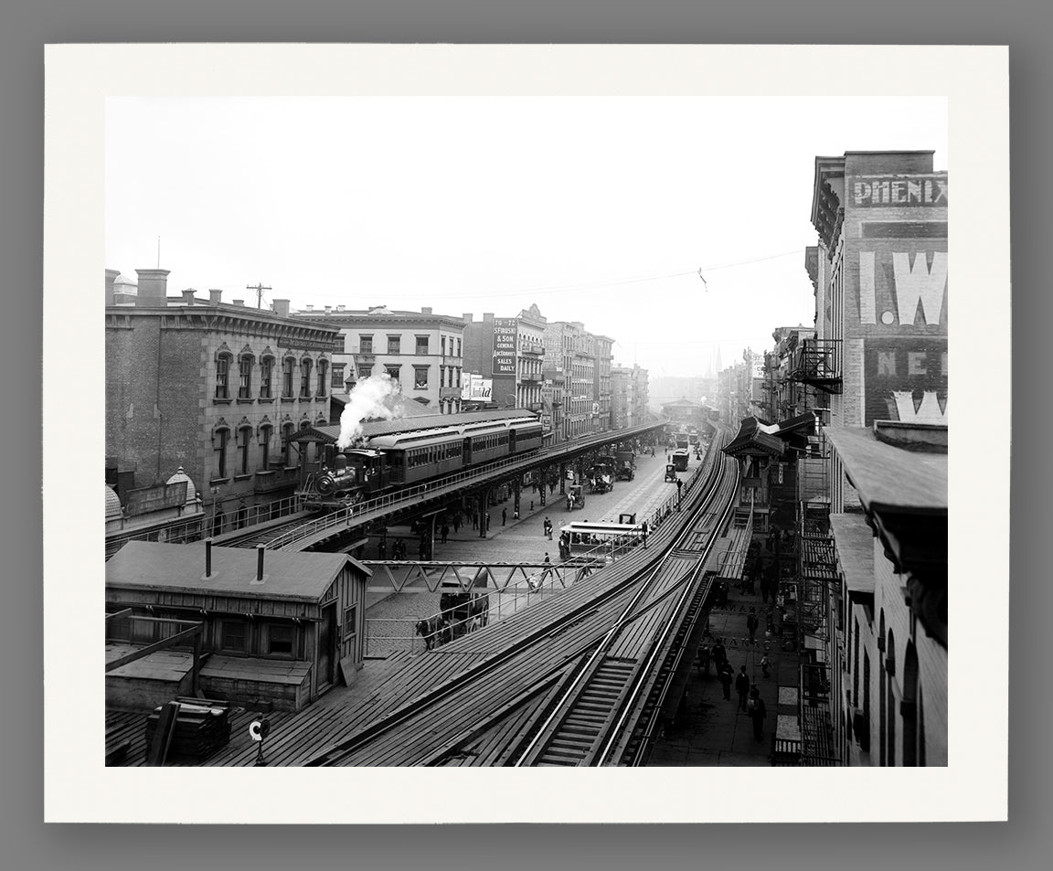 Vintage black and white photograph of a New York City street with train tracks, buildings, and people, printed on paper.