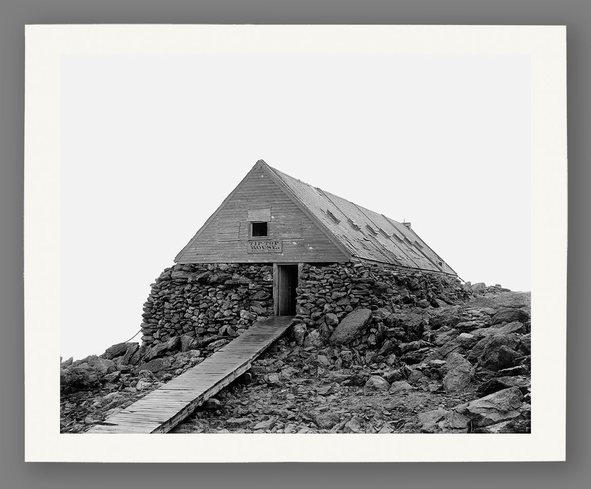 Paper print of a black and white photograph of a stone building on a rocky mountain peak with a wooden staircase.
