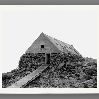 Paper print of a black and white photograph of a stone building on a rocky mountain peak with a wooden staircase.