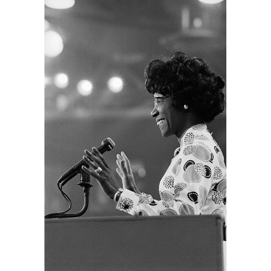 Black and white photo of Shirley Chisholm speaking into a microphone at a podium.
