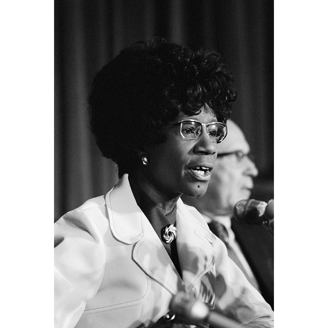 A vintage, black and white photo of Shirley Chisholm speaking into a microphone