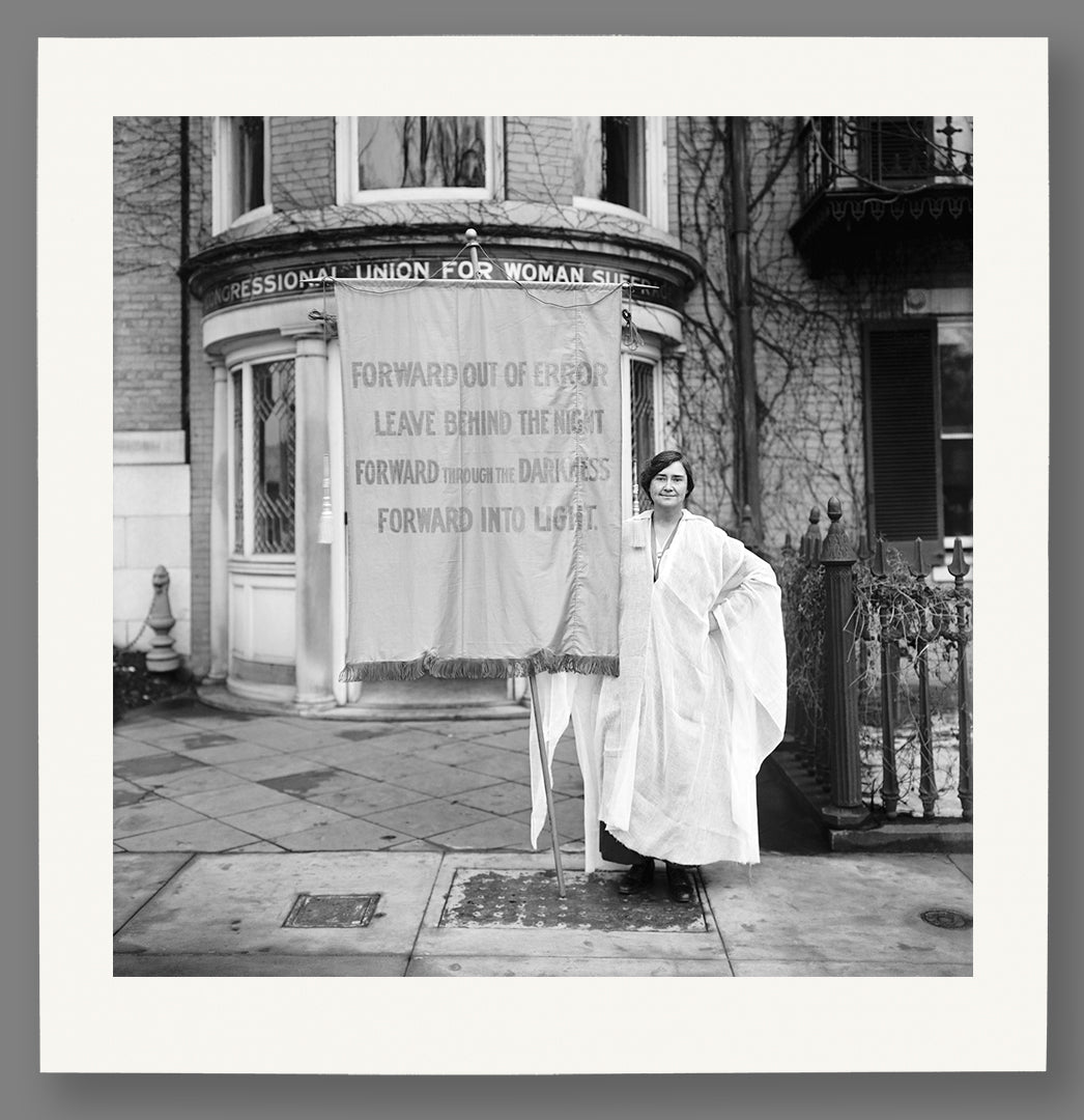 Paper print of a vintage photo of a woman standing in front of a banner for the Suffragette movement outside a building.