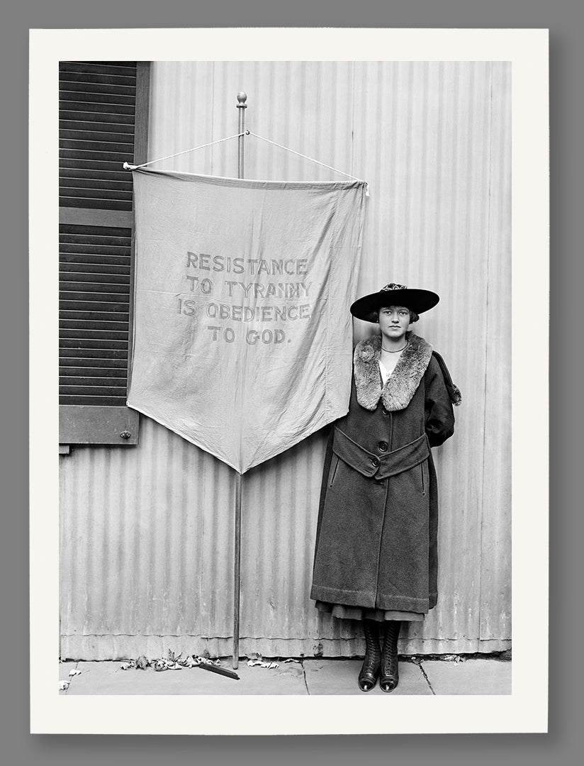 A paper print of a photo of a woman standing next to a banner with text, wearing a coat and hat, in front of a building.