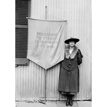 Suffragette standing next to a banner with text, wearing a coat and hat, in front of a corrugated metal wall.