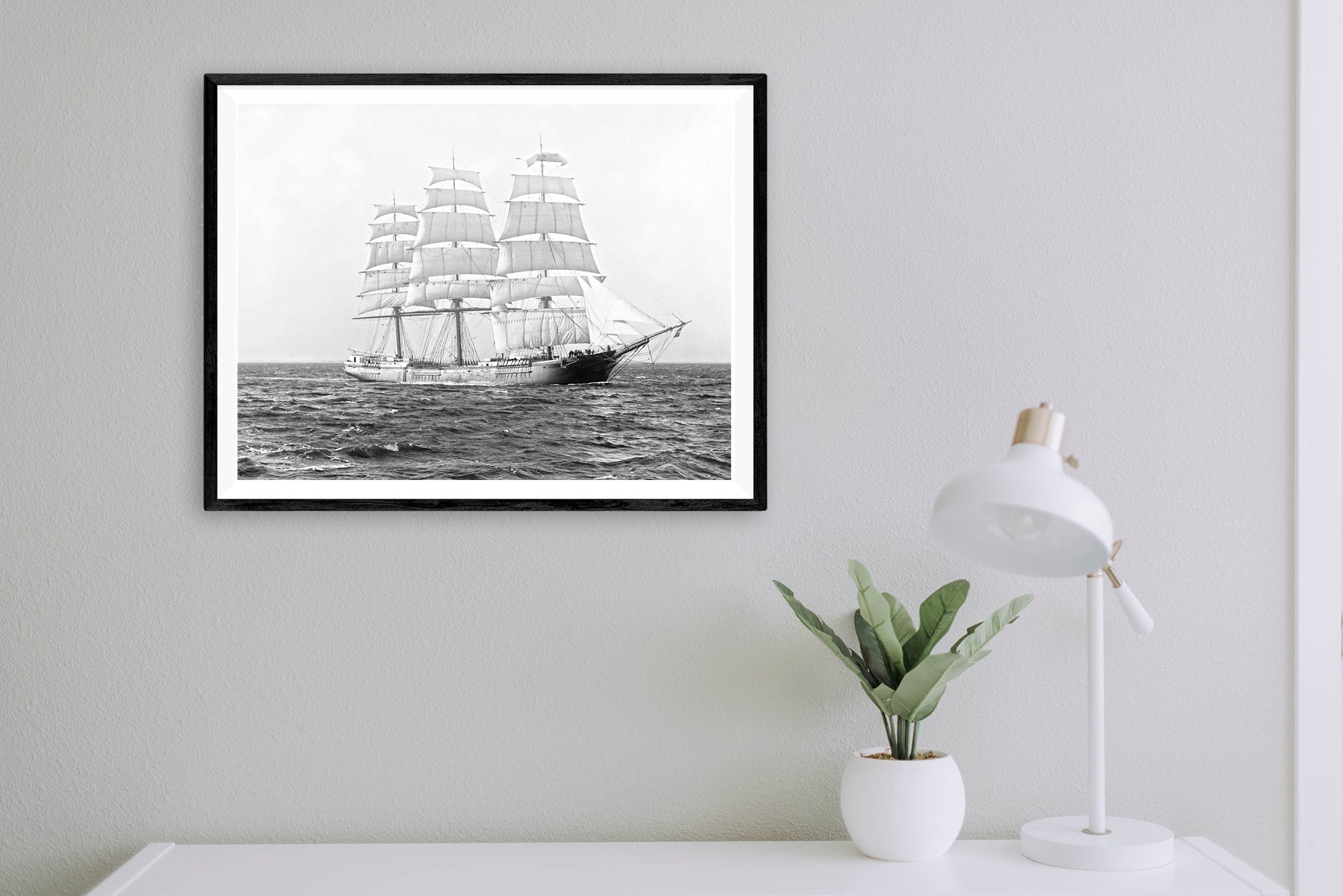 A framed paper print of a vintage photograph of a Clipper Ship hanging above a small white desk