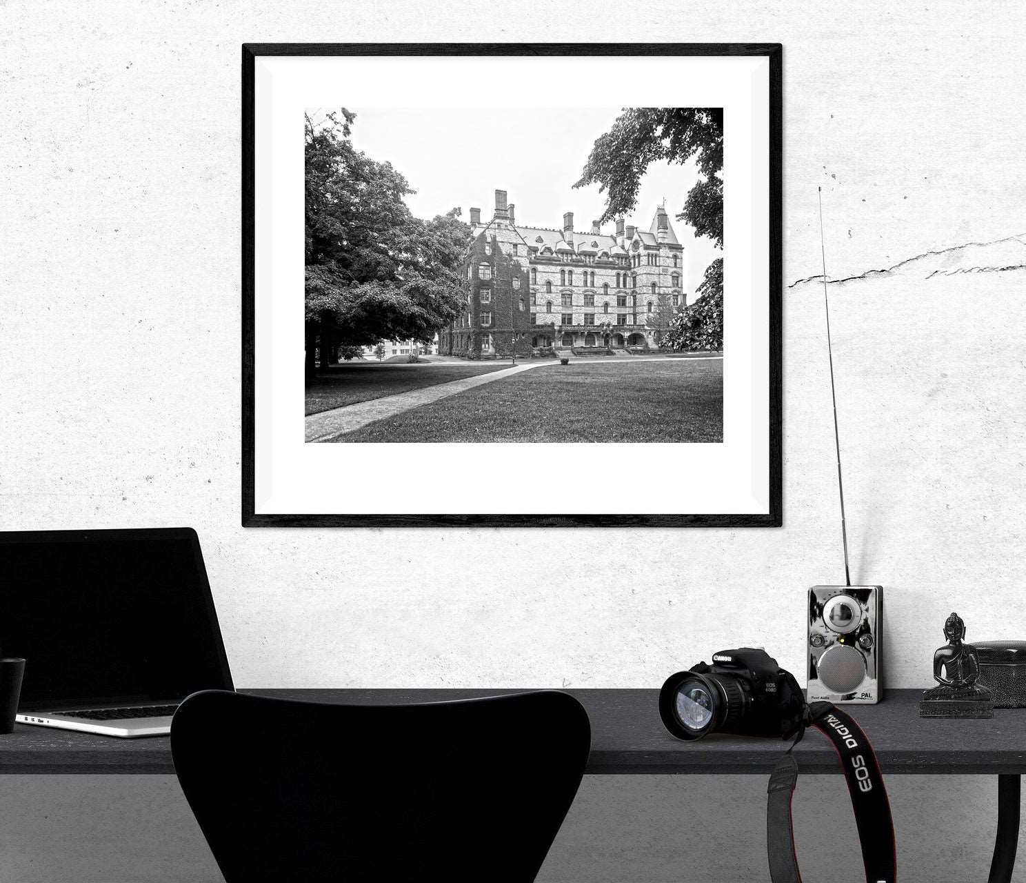 A desk with a laptop and camera, with a framed paper print hanging above featuring a vintage photo of Witherspoon Hall